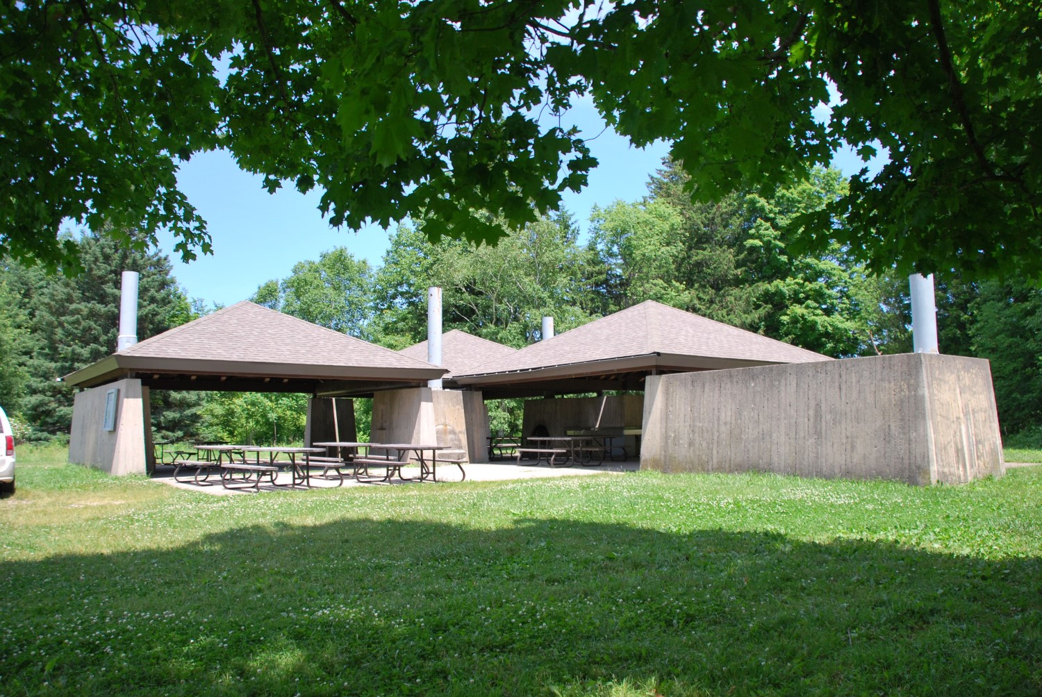 A cement picnic shelter with two pyramid-shaped roofs on top. The shelter is set back in a grassy field.