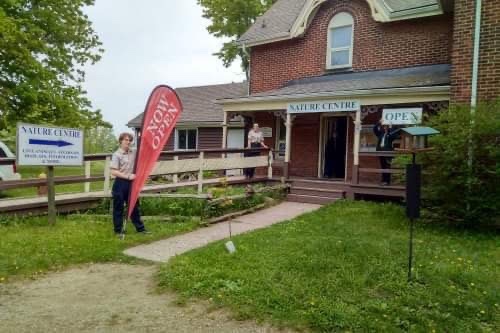 An Ontario Parks staff person wearing their beige and black uniform standing outside the Nature Centre, a red brick building.