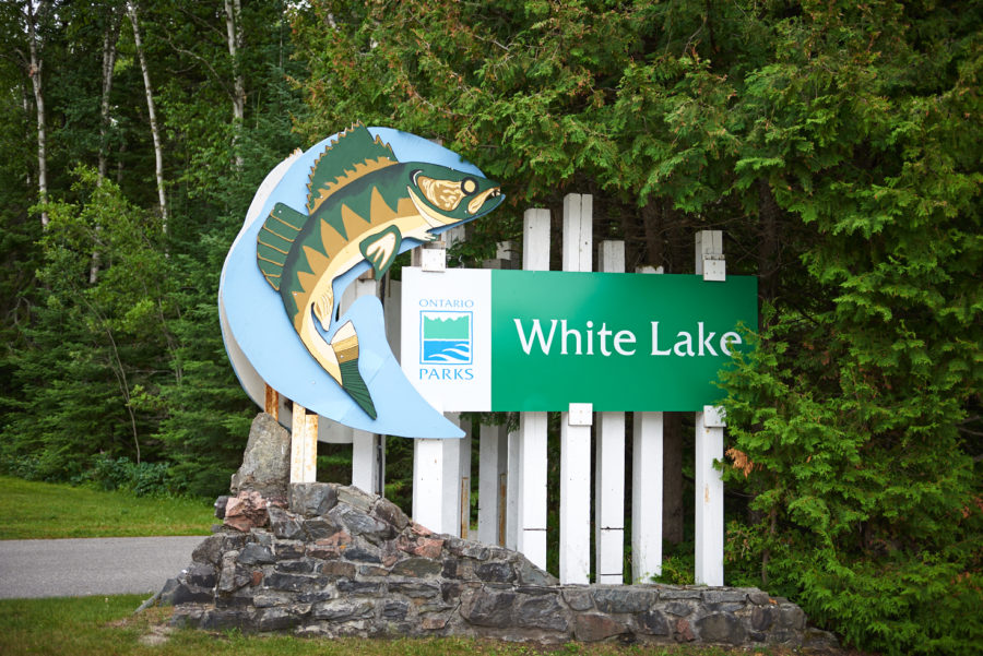 Entrance sign at White Lake. Multiple white poles holding the name sign and to the left a large fish art piece. The park which Mitch is fishing at.