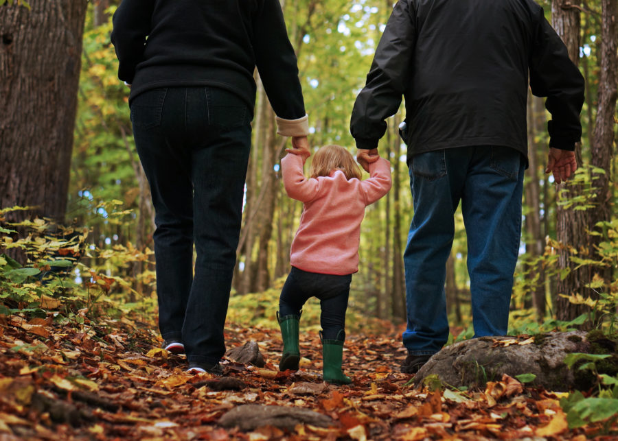 Une famille de trois marche sur des feuilles d’automne le long d’un sentier. L’enfant est au milieu.
