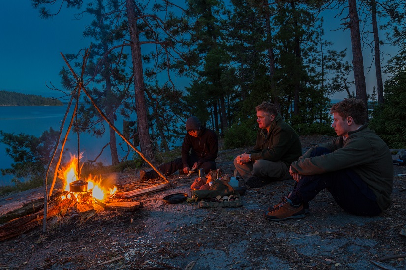 Three men sit around campfire on rock