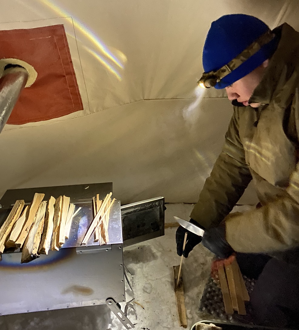 Man in a hot tent adds wood to a stove