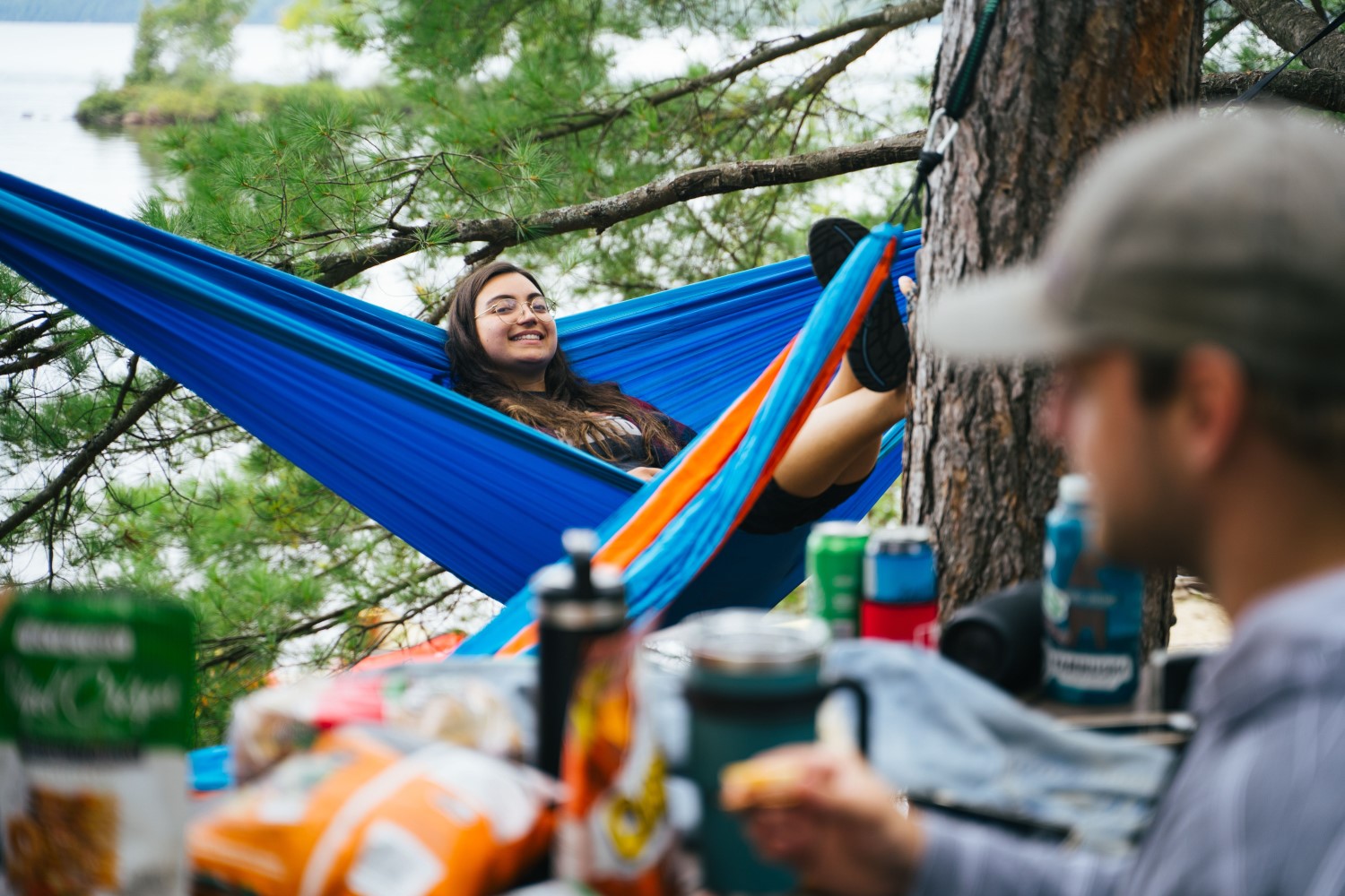 A person lying in a hammock, smiling at their camping companion who is sitting at the picnic table in the foreground.