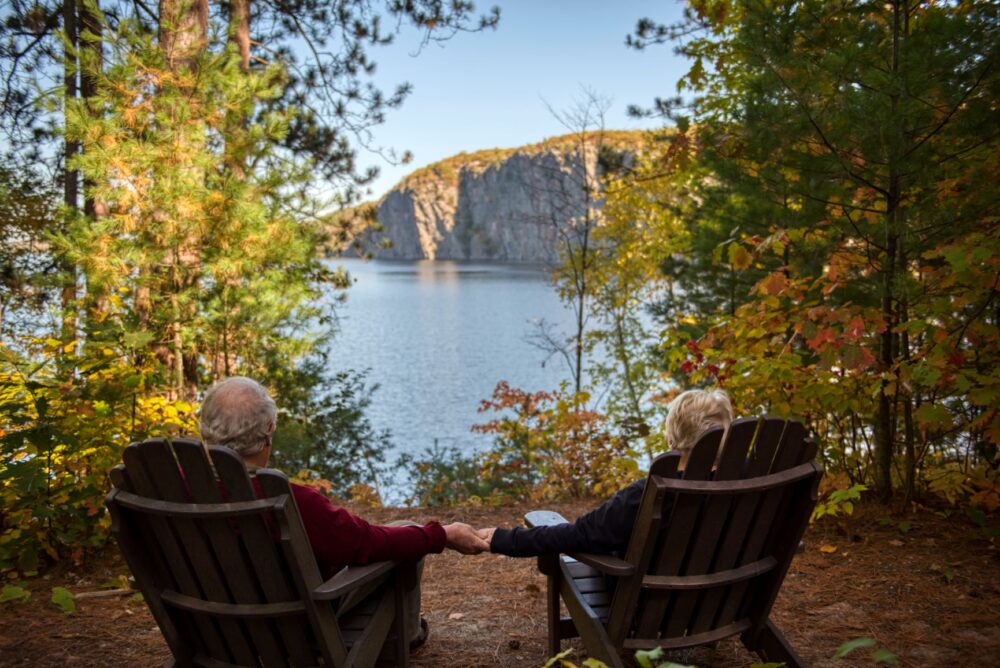 Senior citizens sitting in wooden chairs holding hands overlooking fall view with lake and cliffs