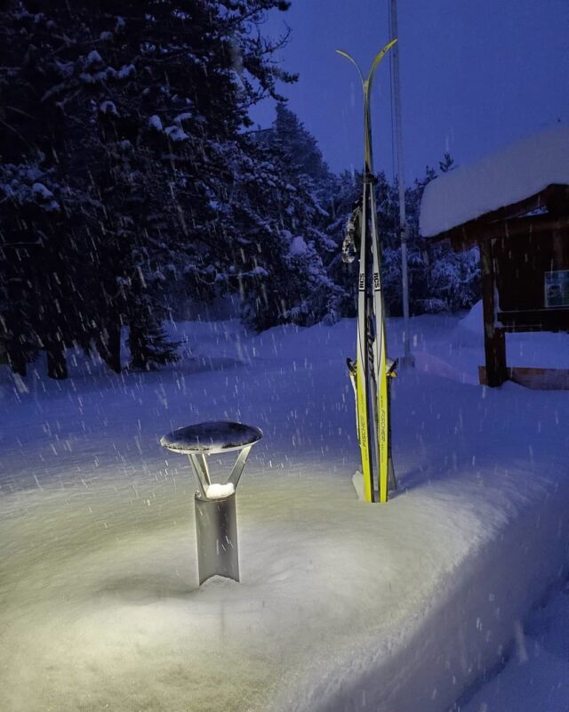 A shielded light that lights up the snow with a pair of cross-country skis sticking out of it. 