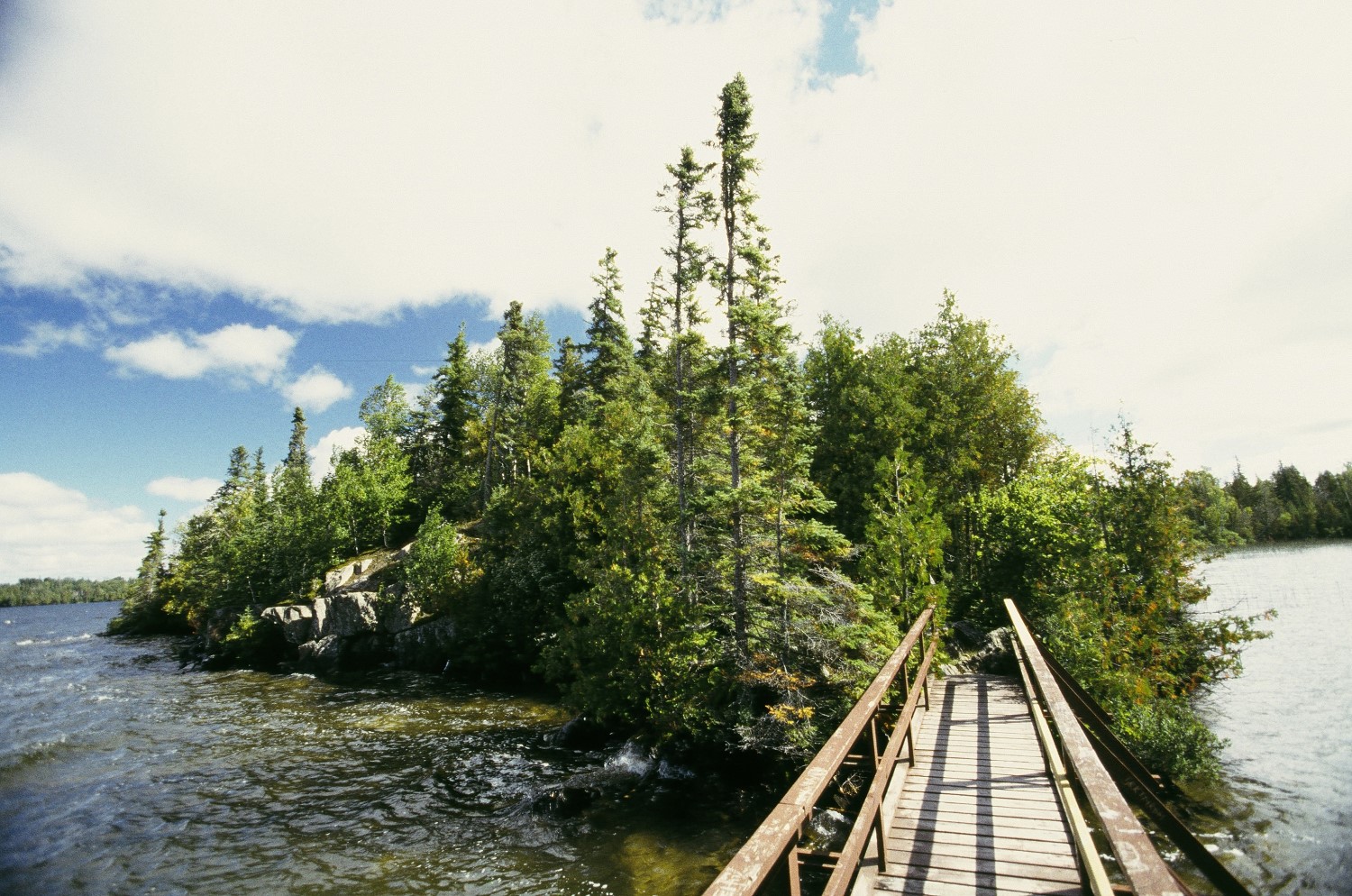 Un étroit pont piétonnier en bois sur l'eau menant à une petite île boisée sous un ciel ensoleillé.