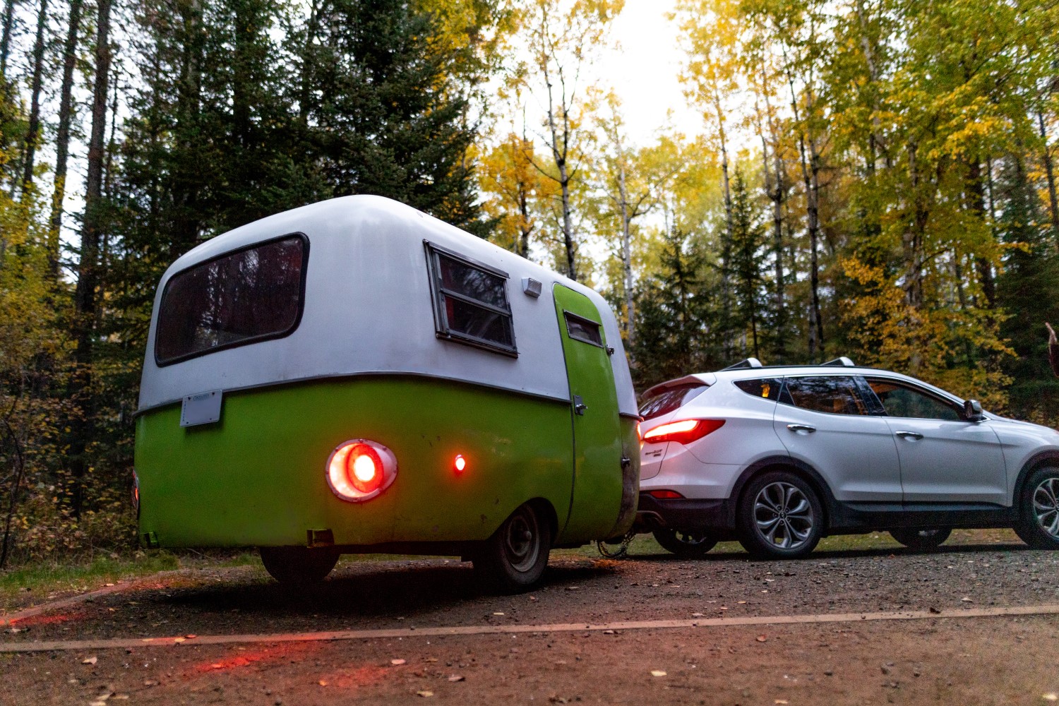 Une petite roulotte verte et blanche attachée à un VUS argenté sur le stationnement d'un terrain de camping.