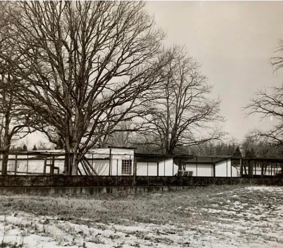 Historical photo in black and white, showing a series of low, one-story buildings and fenced areas. Image was taken in winter: there are some large leafless deciduous trees surrounding the buildings, and there is an open field with snow in the foreground.