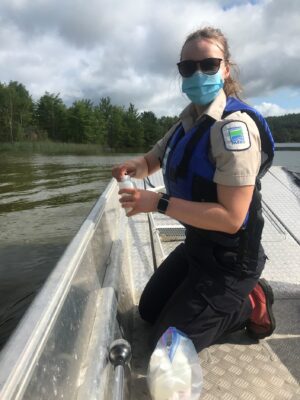 Woman in a boat, wearing an Ontario Parks uniform and life jacket, holding a water sample