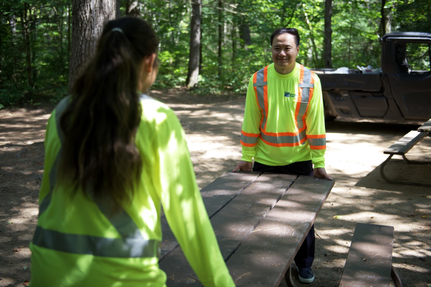 Two Ontario Parks staff members wearing neon yellow high visibility shirts. Together, they are lifting and moving a wooden picnic table.
