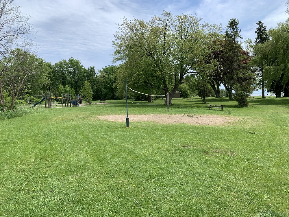 A green space featuring a beach volleyball court, kids' play structures, and a picnic table. The area is hemmed in by tall trees.