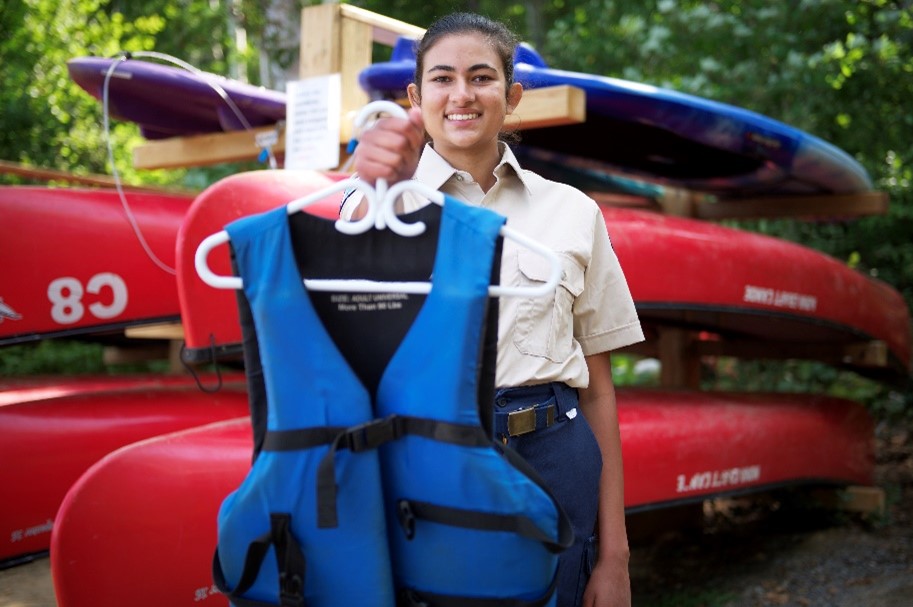 An Ontario Parks staff person holding out a blue PFD on a hanger and smiling. They are standing in front of a kayak rack in the forest.