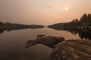 A hazy sky at sunset. The sun is setting over a lake. The sky is a dusty pink. There is a forest off in the distance.