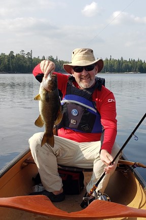 A person sitting in the front of a canoe, facing the camera and smiling, holding a fishing pole in one hand and a fish in the other hand.