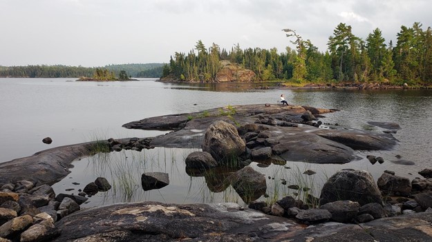 A rockface jutting up through a lake. A person sits on the rock in the distance. Further back, there is a forest on the shoreline across from the rock.