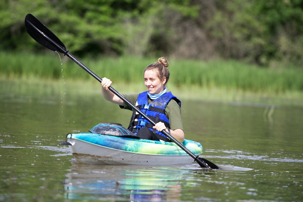 Une personne assise dans un kayak bleu sur l'eau. Elle porte un vêtement de flottaison individuel bleu royal et tient une pagaie à deux mains, avec un côté de la pagaie plongé dans l'eau. Elle sourit et regarde au loin, hors caméra.