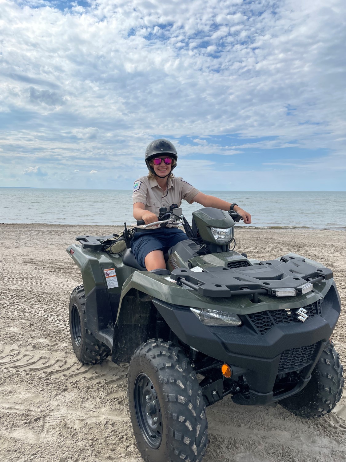 A Park employee wearing a helmet and sunglasses, sitting on an ATV with both hands on the handles. The ATV is parked on a beach with the water and a blue sky behind it.