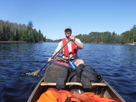 A person sitting at the back of a packed canoe, wearing a PFD and paddling in a lake.