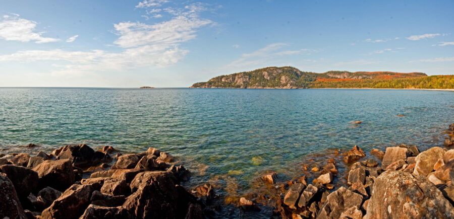 A panoramic view of Lake Superior on a sunny day with clear turquoise water and a rocky shore.