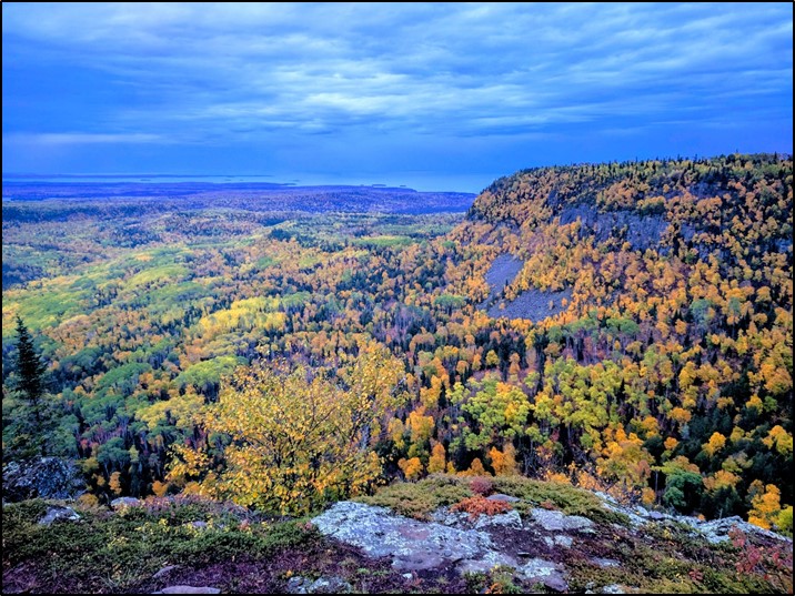 A rock face overlooking a forest changing colour in the fall, with trees that are dark green, light green gold and orange.