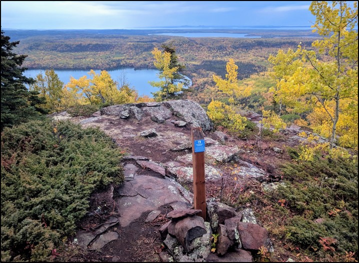 Rocky outlook with a sign post, overlooking a bay surrounded by thick forest.