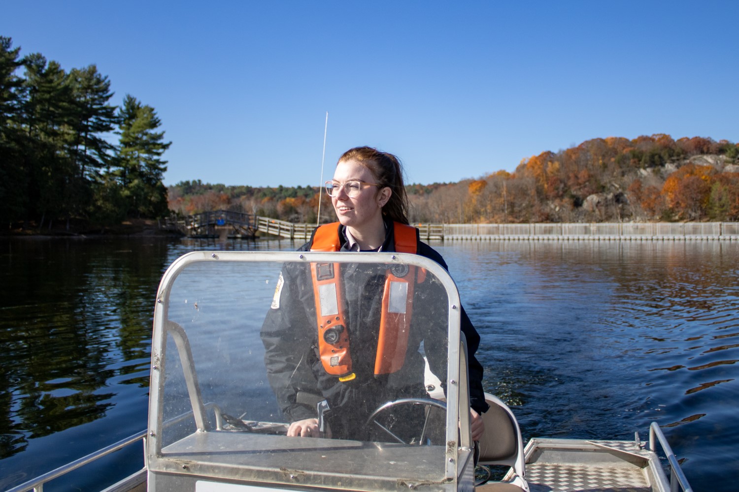 Une personne portant un VFI conduisant un petit bateau à moteur dans un lac, avec une forêt aux couleurs d'automne derrière elle.