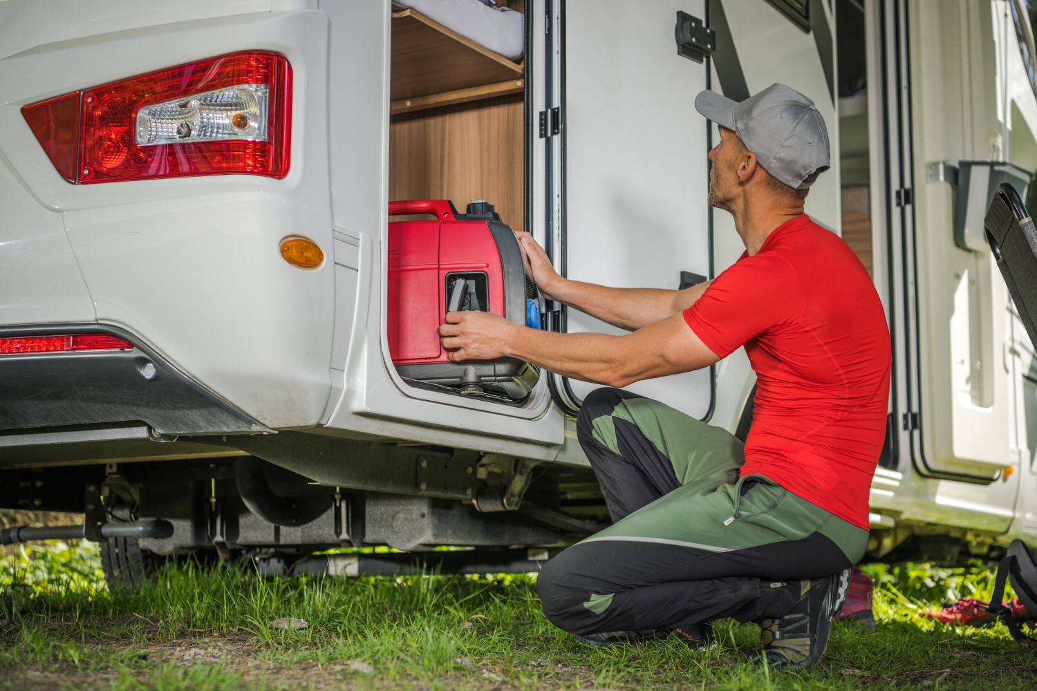 A person crouching on the ground, removing a gas generator from its storage cupboard at the back end of an RV