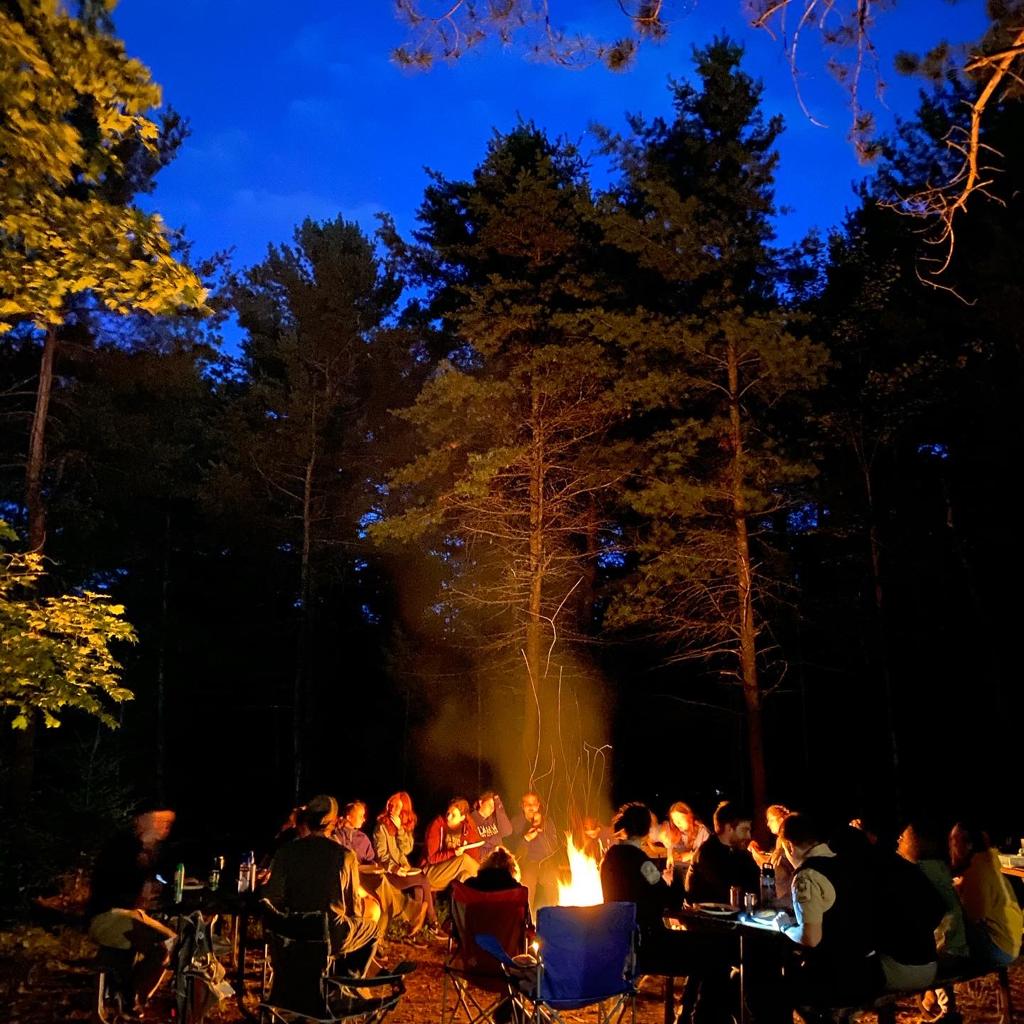 A large group of people in camping chairs circled around a campfire in a forest clearing at nightfall.