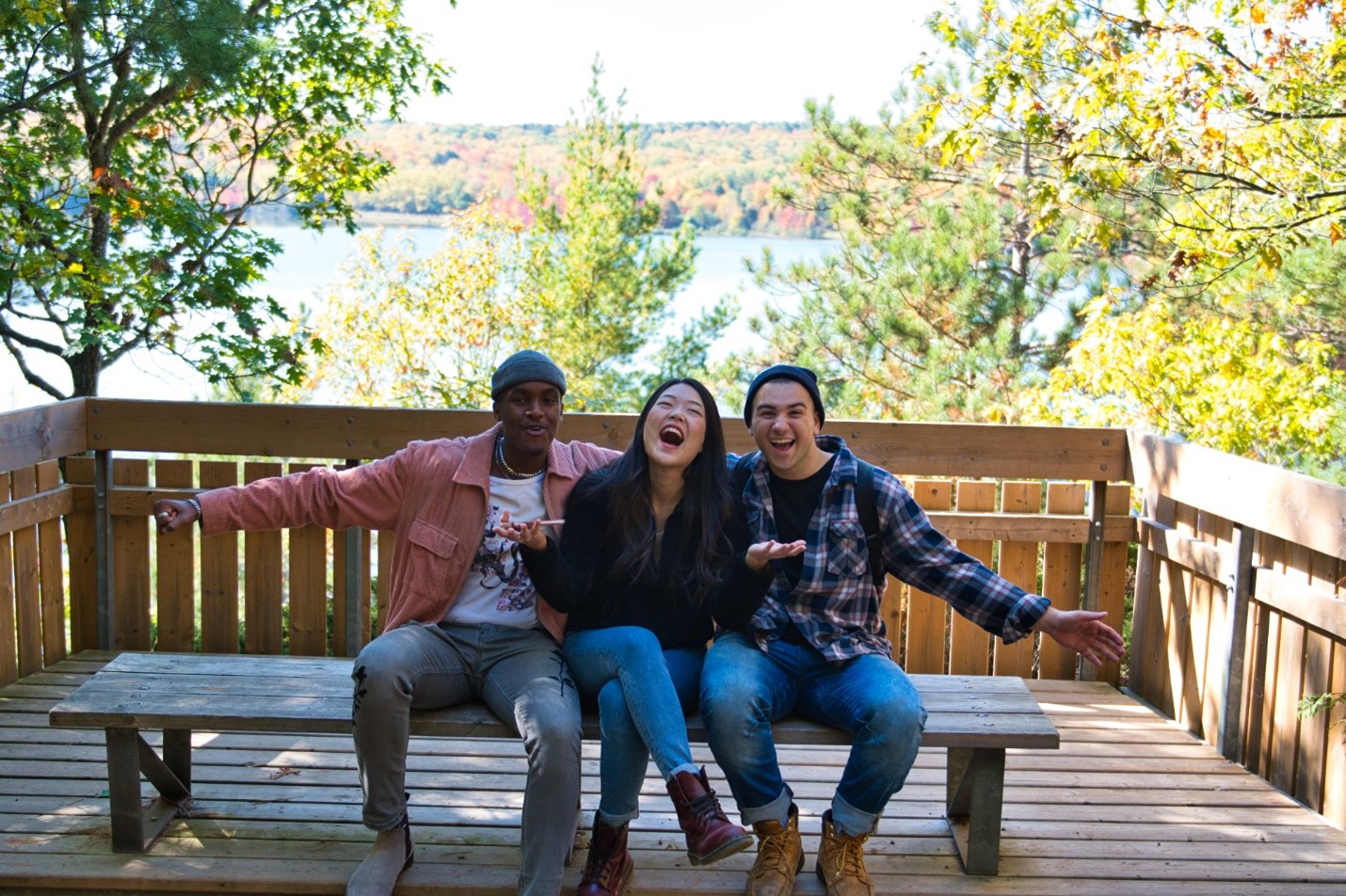 Three young adults sitting on a bench in a forest, smiling widely at the camera with their arms outstretched