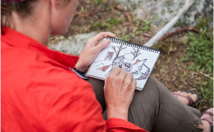 Au-dessus de l’épaule d’une personne portant une chemise rouge, en train de dessiner dans le bois