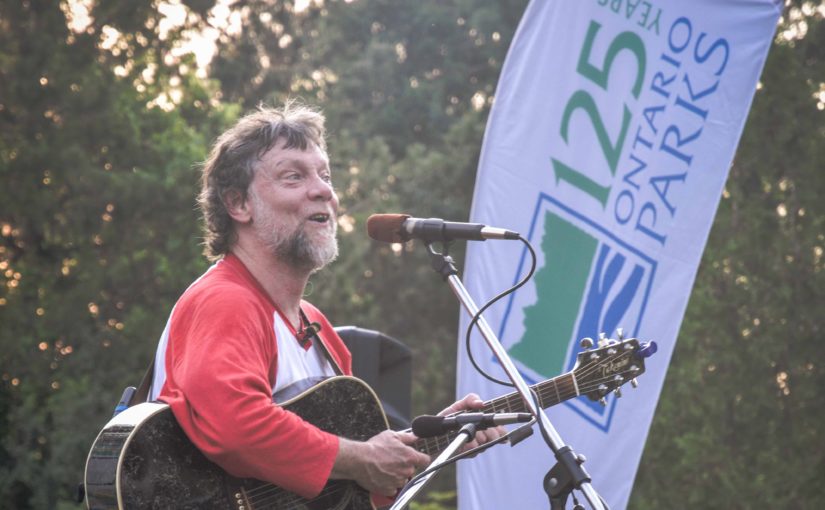 Chanteur portant un chandail rouge et blanc et jouant de la guitare, avec la bannière de Parcs Ontario en arrière-plan.