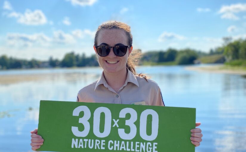 Une personne portant des lunettes de soleil se tient devant un lac. Elle tient un panneau vert rectangulaire sur lequel on peut lire « 30 x 30 Nature Challenge » (Défi dans la nature 30 x 30).