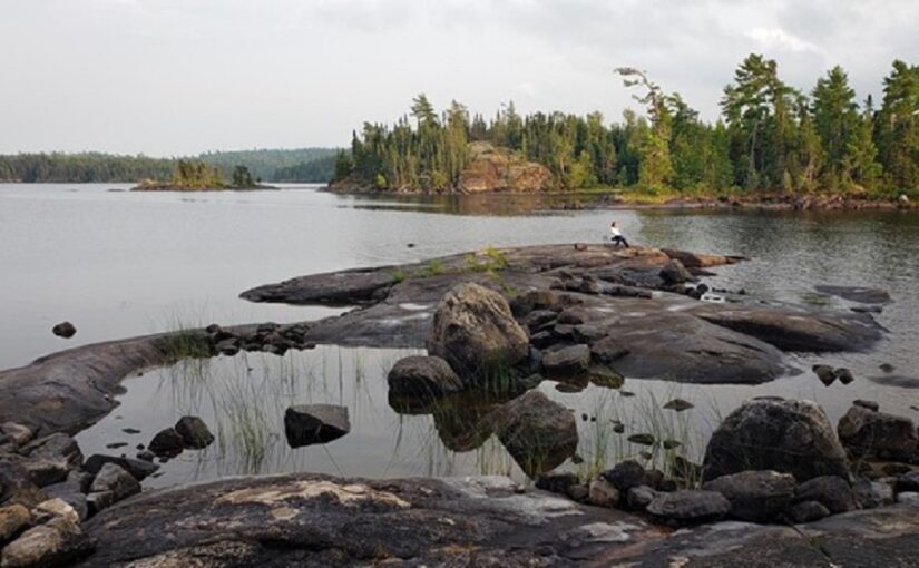 Une paroi rocheuse en saillie dans un lac. Une personne est assise sur le rocher au loin. Plus loin, on aperçoit une forêt sur la rive, en face du rocher.