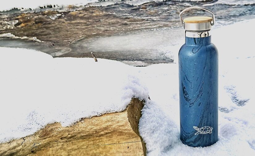 A Chilly Moose blue water bottle on a patch of snow beside a stream.