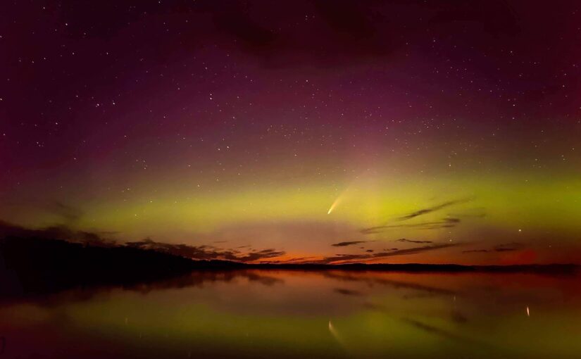 A purple night sky with shimmering bands of yellow and orange northern lights on the horizon. The colours reflect on a still lake below.