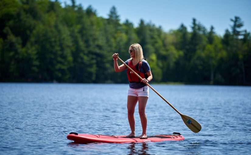 Female with sunglasses on paddleboarding on a blue lake in the summer with a green forest in the background.