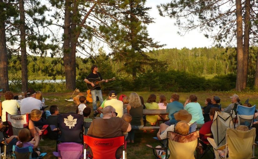 David with a guitar preforming to a crowd in a natural setting, late in the day, in the summer