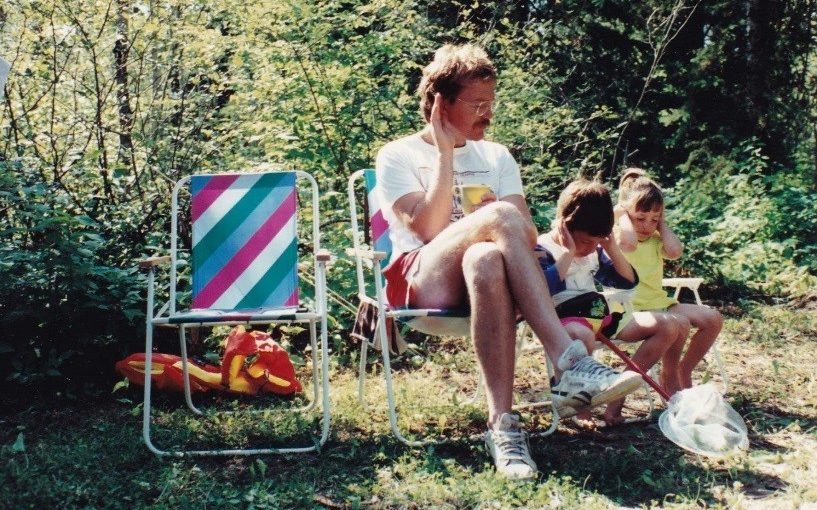 Dad sitting in a colourful lawn chair on teh edge of a forest with two kids (also in lawn chairs), all with their hands to their ears.