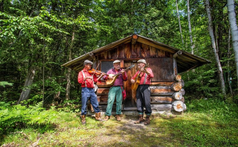 Three red flanel shirt clad men with caps and instruments posing in front of a log cabin in the woods