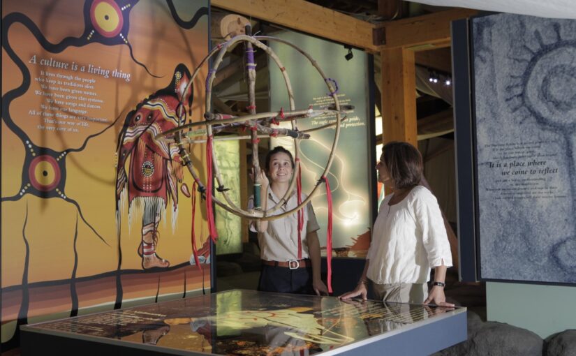 An Ontario Parks staff person looks at a 3D hoop medicine wheel with a visitor in the Petroglyphs visitor centre. On the wall behind the wheel is a large Indigenous painting of a person in red clothing on an orange backdrop.