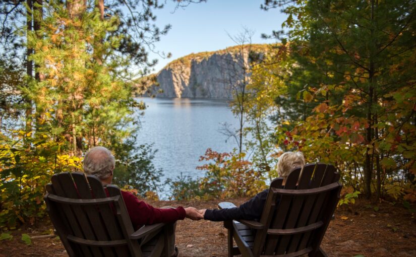A couple sitting in Muskoka chairs holding hands while looking out at the lake