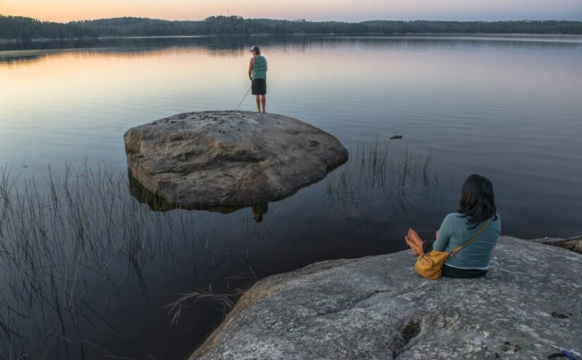 A person sitting on a rock on the shoreline watching another person fish on a rock further into the water.
